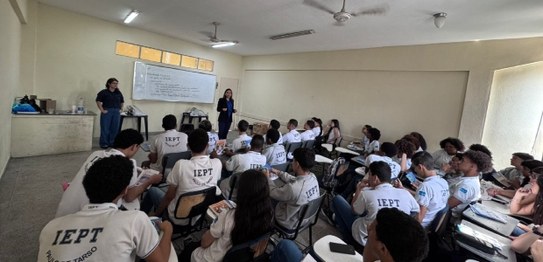 Auditório com cortina vermelha ao fundo, onde um homem está à frente, conduzindo uma apresentaçã...