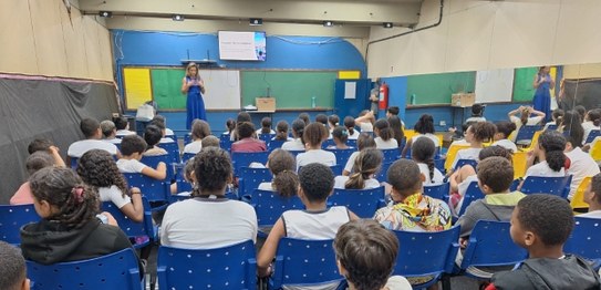 Auditório com cortina vermelha ao fundo, onde um homem está à frente, conduzindo uma apresentaçã...