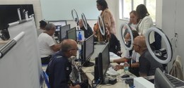 Guichês de atendimento, com fileira de pessoas sentadas; espaço com computadores e equipamentos ...