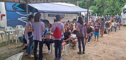 Descrição da imagem principal: Em pé, quatro voluntárias da Justiça Itinerante atendem crianças,...