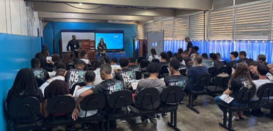 Um palestrante fala para jovens sentados em cadeiras azuis em um auditório simples, com bandeira...