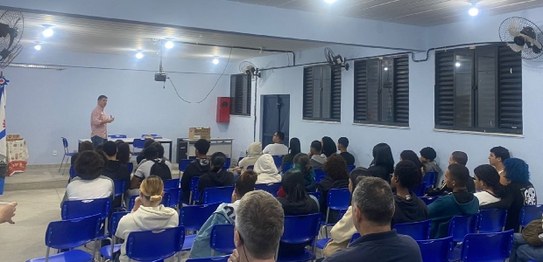 Um palestrante fala para jovens sentados em cadeiras azuis em um auditório simples, com bandeira...