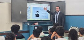 Magistrado apresenta slide sobre os Três Poderes em sala de aula, apontando para a tela enquanto...