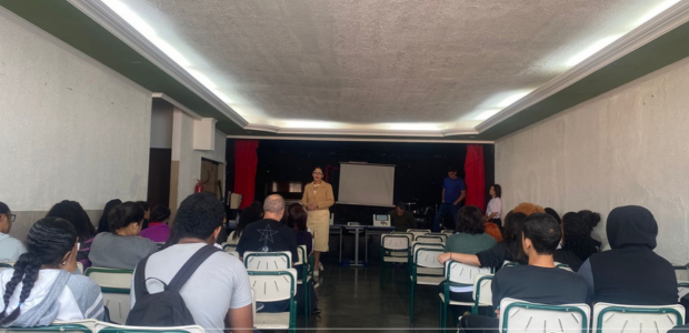 A imagem mostra uma sala ampla, com paredes claras e teto branco, onde várias pessoas estão sent...