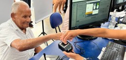 A imagem mostra um idoso de camisa listrada branca realizando um procedimento de biometria. Ele ...