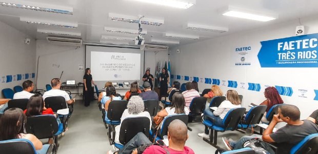 A imagem mostra uma sala de aula da Faetec Três Rios, com várias pessoas sentadas em cadeiras es...