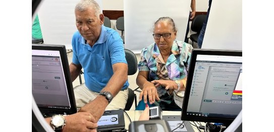A imagem mostra um casal idoso sendo atendido em um local que parece ser um mutirão de emissão d...