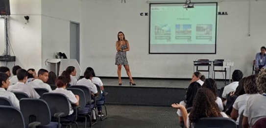 A imagem mostra um ambiente de sala de aula com um quadro branco ao fundo e estudantes sentados....