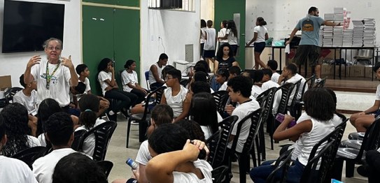 A imagem mostra um grupo de crianças sentadas no chão, vestindo uniformes escolares brancos, ass...
