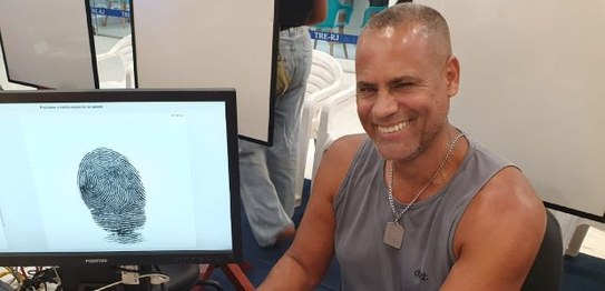 Na imagem, vemos um homem sentado em frente a um monitor de computador. Ele sorri para a câmera....