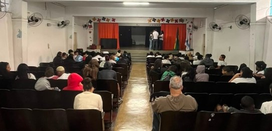 Em uma sala ampla e iluminada, quatro palestrantes de pé ao lado de bandeiras apresentam uma ati...