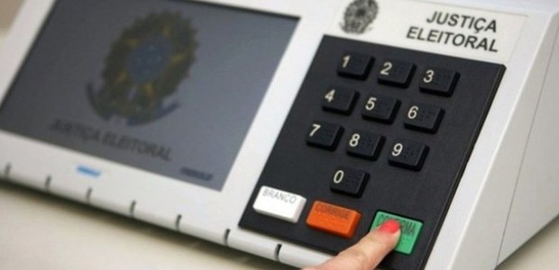 No Brasil, só é possível votar presencialmente foto em close da urna eletrônica, onde temos um dedo que aperta o botão “Confirma”