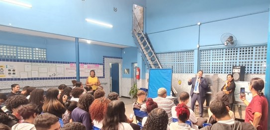 Numa sala de aula, sentados, estudantes assistem à palestra do juiz eleitoral. Vestindo terno pr...