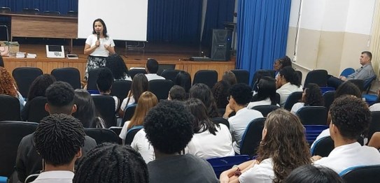 Numa sala, sentados, estudantes assistem à palestra da juíza eleitoral. Ela está de pé na frente...