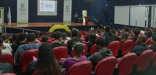 Numa sala, sentados, estudantes assistem à palestra da juíza eleitoral. Ela está de pé na frente...