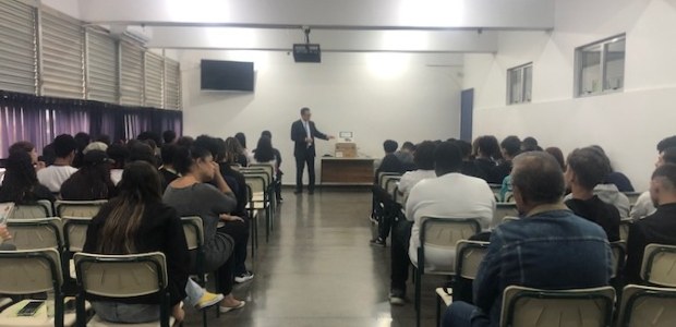 Numa sala de aula, em primeiro plano, sentadas(os) e de costas, estudantes assistem à palestra do juiz eleitoral. Ele está de pé, na frente das(os) estudantes, falando e gesticulando, atrás dele, pendurada na parede, uma televisão. Do seu lado direito, em cima de uma mesa, uma urna eletrônica. Numa sala de aula, em primeiro plano, sentadas(os) e de costas, estudantes assistem à palestra d...