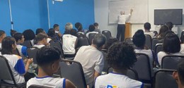 Numa sala de aula, em primeiro plano, sentados e de costas, estudantes da Escola Municipal Aquin...