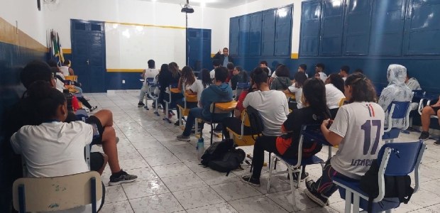 Numa sala de aula, sentados, em primeiro plano e de costas, estudantes assistem à palestra. A foto foi tirada do fundo da sala, e o chefe do cartório está de pé na frente da turma, gesticulando. Numa sala de aula, sentados, em primeiro plano e de costas, estudantes assistem à palestra. A fo...