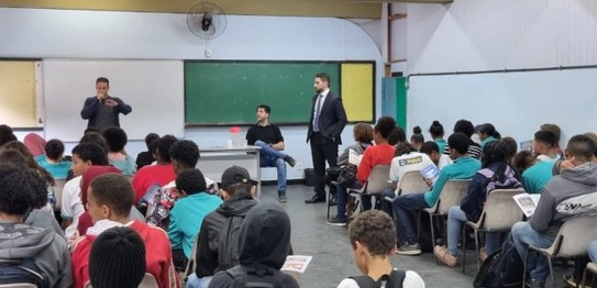Numa sala de aula, sentados, estudantes assistem à palestra do juiz eleitoral. Vestindo terno pr...
