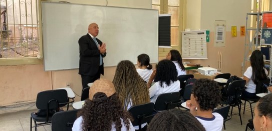 Numa sala de aula, sentados, estudantes assistem à palestra do juiz eleitoral. Vestindo terno pr...