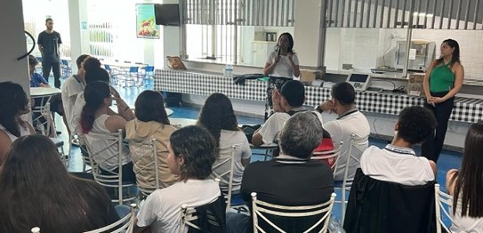 Numa sala, sentados, estudantes assistem à palestra da juíza eleitoral. Ela está de pé na frente...