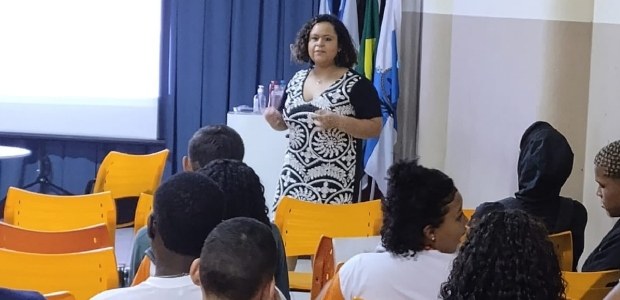 Numa sala, sentados, estudantes assistem à palestra da juíza eleitoral. Ela está de pé na frente da turma, gesticulando e falando. Atrás dela, numa base, algumas bandeiras, como a do Brasil e a do estado do Rio de Janeiro. Numa sala, sentados, estudantes assistem à palestra da juíza eleitoral. Ela está de pé na frente...