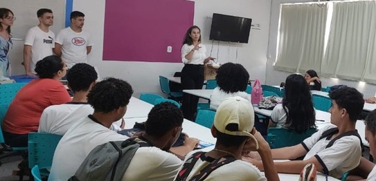Numa sala, sentados, estudantes assistem à palestra da juíza eleitoral. Ela está de pé na frente...