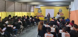 Numa sala, sentados, estudantes assistem à palestra da juíza eleitoral. Vestindo um vestido long...