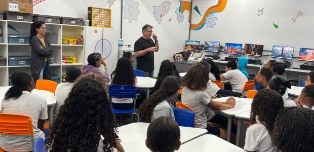 Em uma sala de aula decorada com desenhos nas paredes, um palestrante fala para estudantes senta...