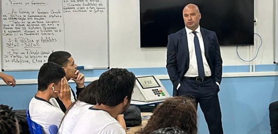 A imagem mostra um ambiente de sala de aula com um quadro branco ao fundo e estudantes sentados....