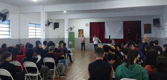 Em uma sala ampla e iluminada, quatro palestrantes de pé ao lado de bandeiras apresentam uma ati...