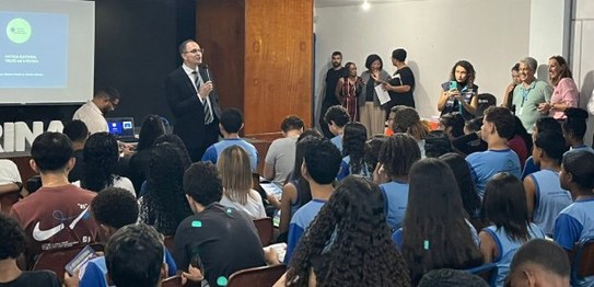 Dois homens falam para um grupo de crianças em uma sala de aula. Uma urna eletrônica está posici...