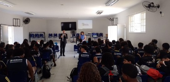 Sala de aula com estudantes sentados em cadeiras azuis assistindo a uma apresentação conduzida p...