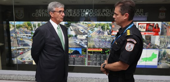 Dois homens conversam. Ao fundo, telas mostram imagens de câmeras de segurança da cidade