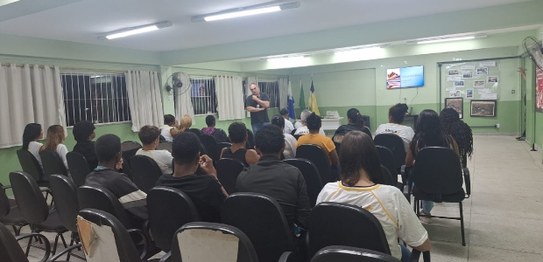 Sala de aula com estudantes sentados em cadeiras azuis assistindo a uma apresentação conduzida p...