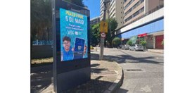 Uma rua com prédios altos, carros estacionados e comércio. Em primeiro plano, há um painel digit...
