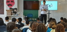 A foto mostra um homem em pé dando uma palestra para um grupo de estudantes em uma sala de aula....