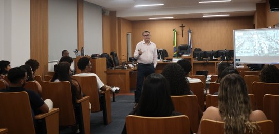 A imagem mostra um homem palestrando para um grupo de jovens sentados em uma sala com computador...