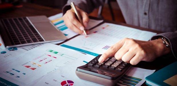 Pessoa trabalhando em uma mesa de escritório repleta de documentos financeiros. Com uma das mãos...