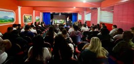 Auditório com várias pessoas sentadas assistindo a uma apresentação no palco; dois apresentadore...