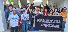 Grupo posa para foto em área coberta, em frente a um cartaz preto com a frase "#PARTIU VOTAR" em...