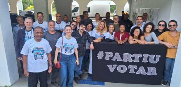 Grupo posa para foto em área coberta, em frente a um cartaz preto com a frase "#PARTIU VOTAR" em...