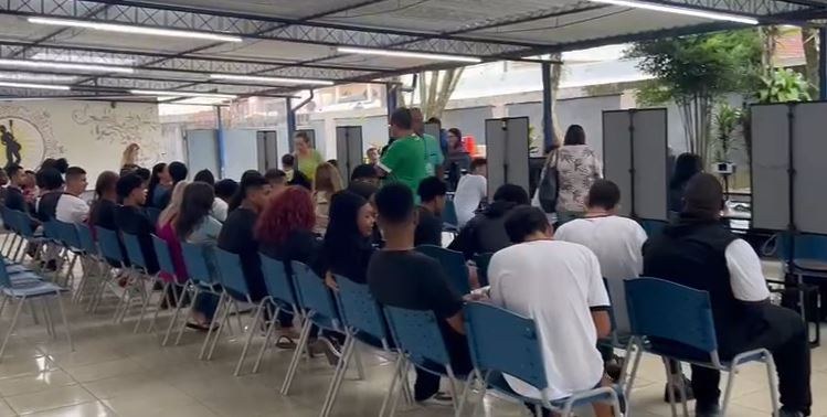 #PartiuVotar tem recorde de atendimentos em Petrópolis A imagem mostra um espaço coberto ao ar livre onde várias pessoas estão sentadas em cadeiras azu...
