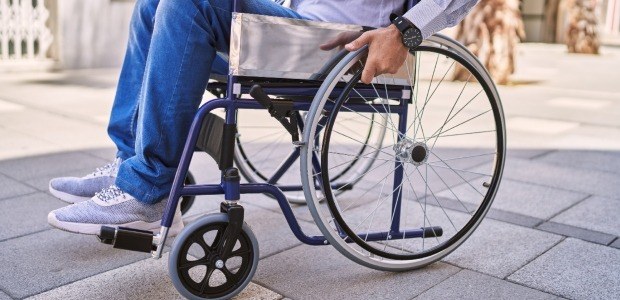 Pessoa sentada em uma cadeira de rodas, vista de lado, movimentando-se em área externa pavimenta...