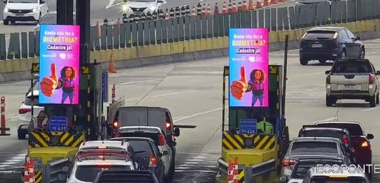 Praça do pedágio da Ecovias Ponte. Em cada guichê, há um painel digital exibindo um aviso sobre ...