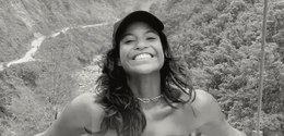Foto posada de Juliana, jovem, sorridente, usando boné escuro e colar, posando ao ar livre em um...