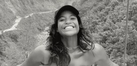 Foto posada de Juliana, jovem, sorridente, usando boné escuro e colar, posando ao ar livre em um...