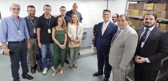 A imagem mostra um grupo de nove pessoas (sete homes e duas mulheres) posando numa sala, onde há...