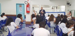 Descrição da imagem: Na sala e de frente para os(as) alunos(as), o juiz apresenta o tema da ação...