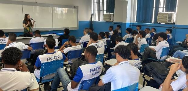 Sala de aula com estudantes uniformizados sentados e atentos, enquanto a palestrante fala ao mic...